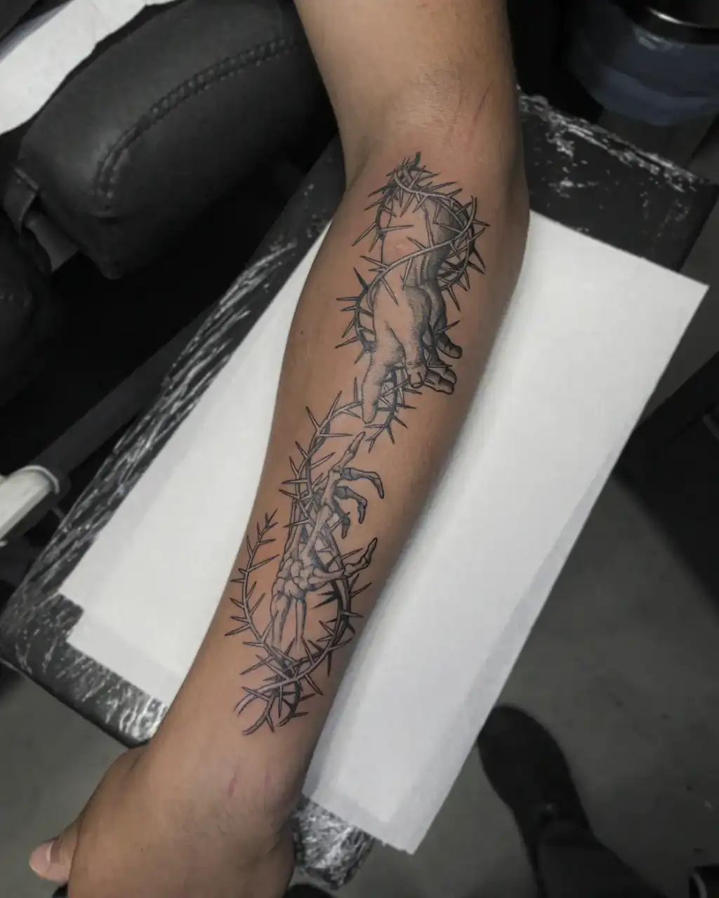 Antebrazo con dos manos, una de ellas esquelética, atrapadas entre ramas de espinas, en black & grey de línea fina.