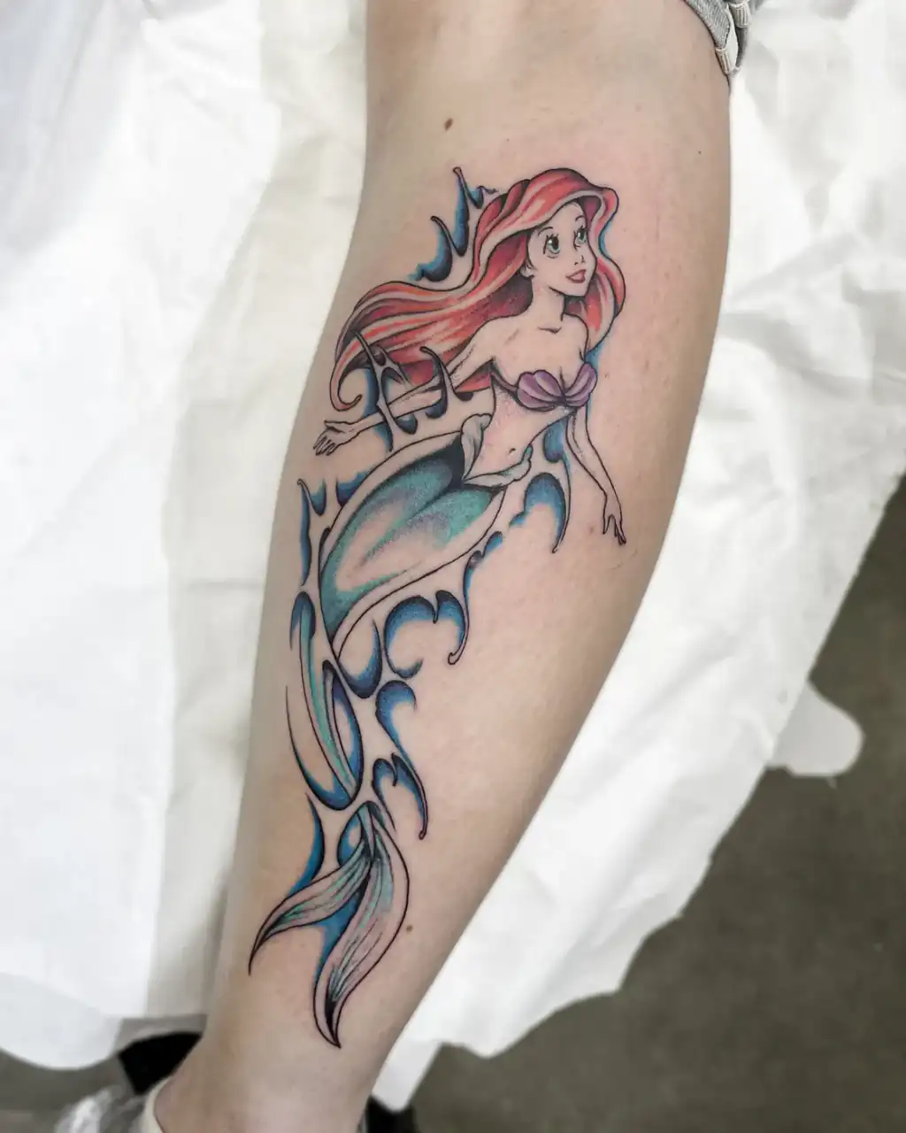 Pantorrilla con una sirena de melena pelirroja y cola verde entre olas azules, tatuada a color.