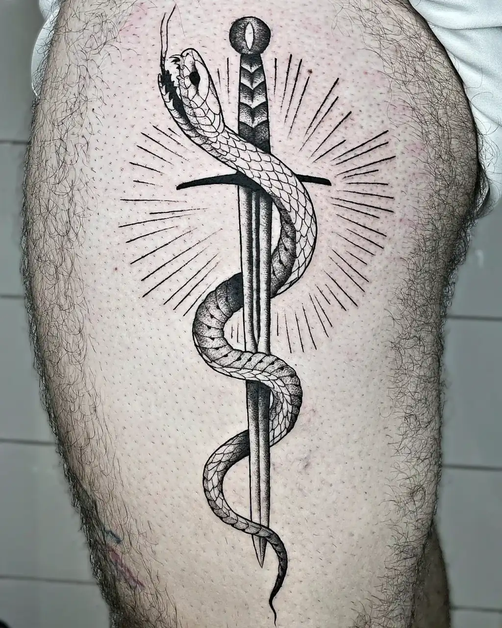 Daga con serpiente y rayos de sol tatuada en el muslo.
