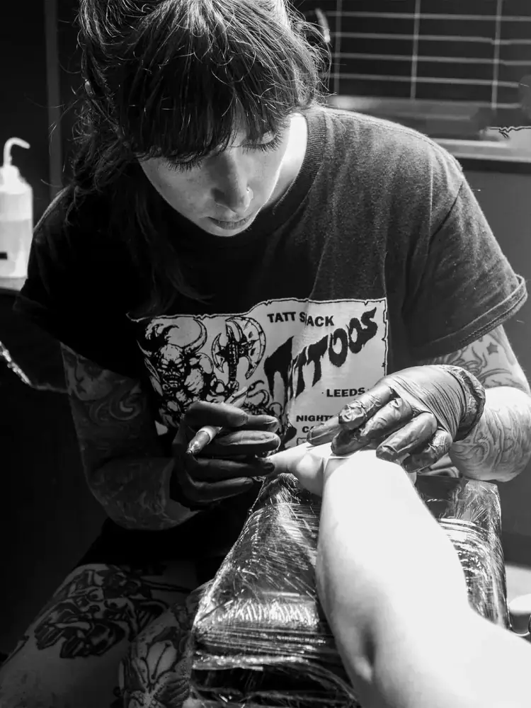 María tatuando con guantes negros el antebrazo de un cliente; camiseta con estampado de tatuaje, en blanco y negro