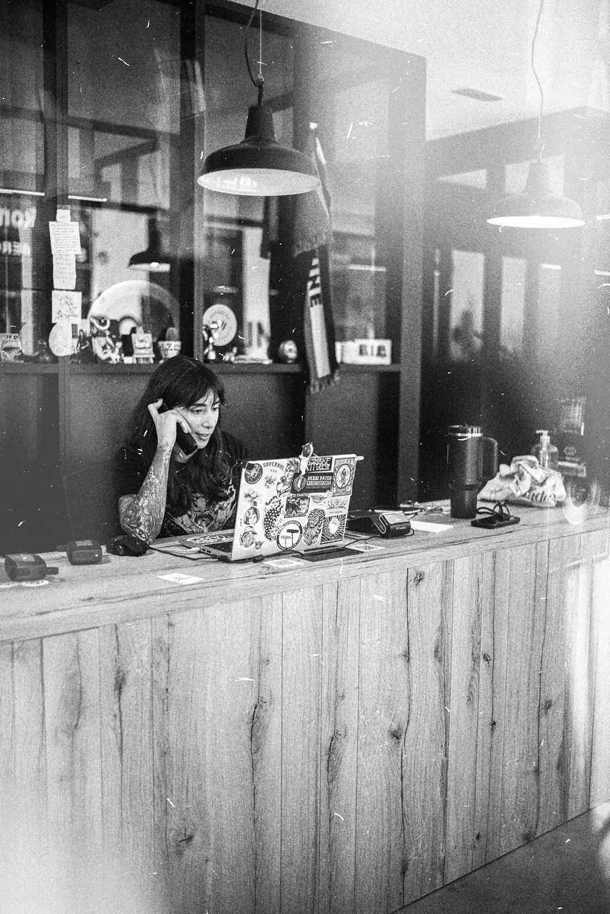 María al teléfono tras el mostrador de madera del estudio, con el portátil forrado de pegatinas. Fotografía en blanco y negro.