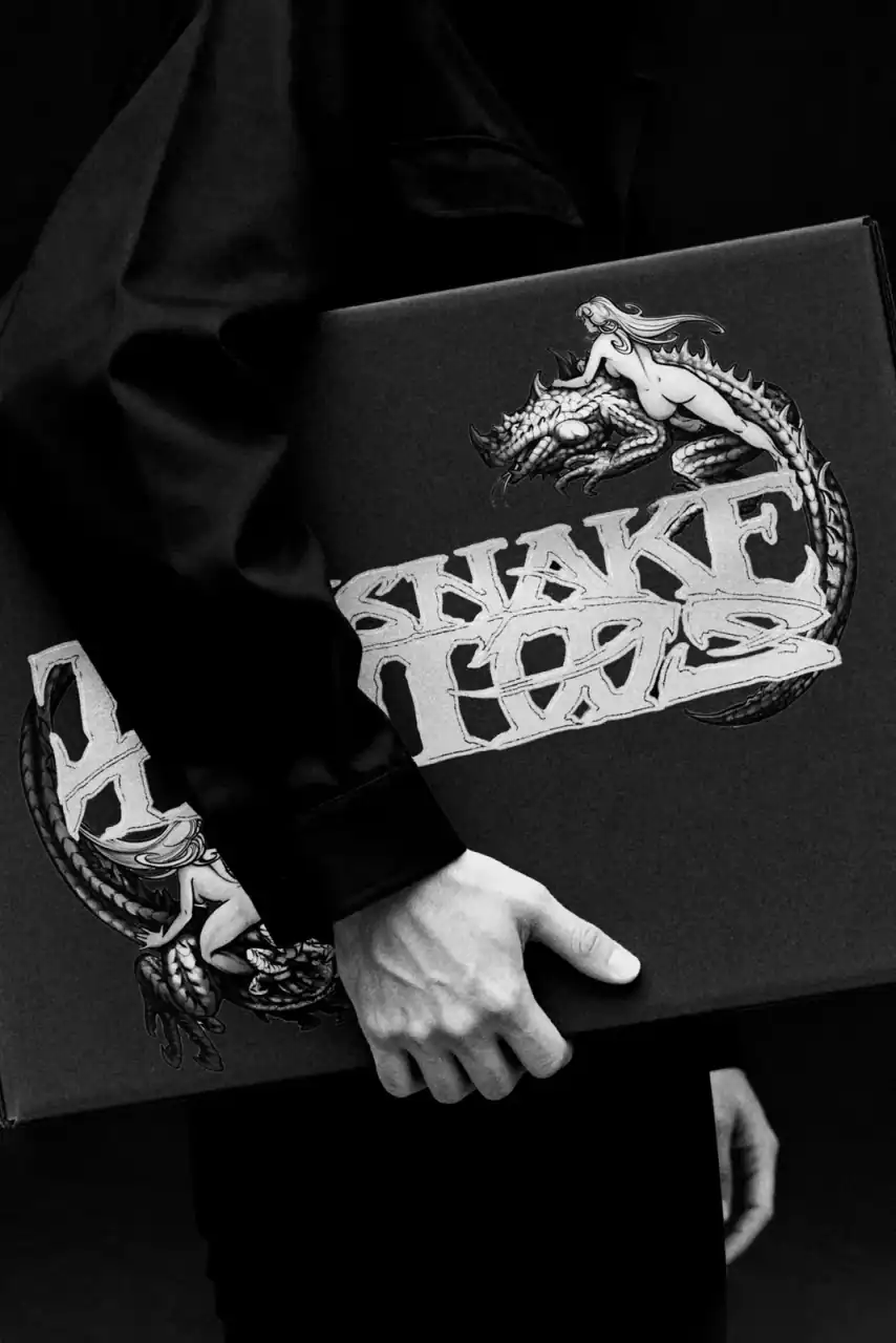 Maqueta de caja negra serigrafiada con el logotipo de InkSnake Tattoo y una serpiente-dragón, sostenida bajo el brazo.